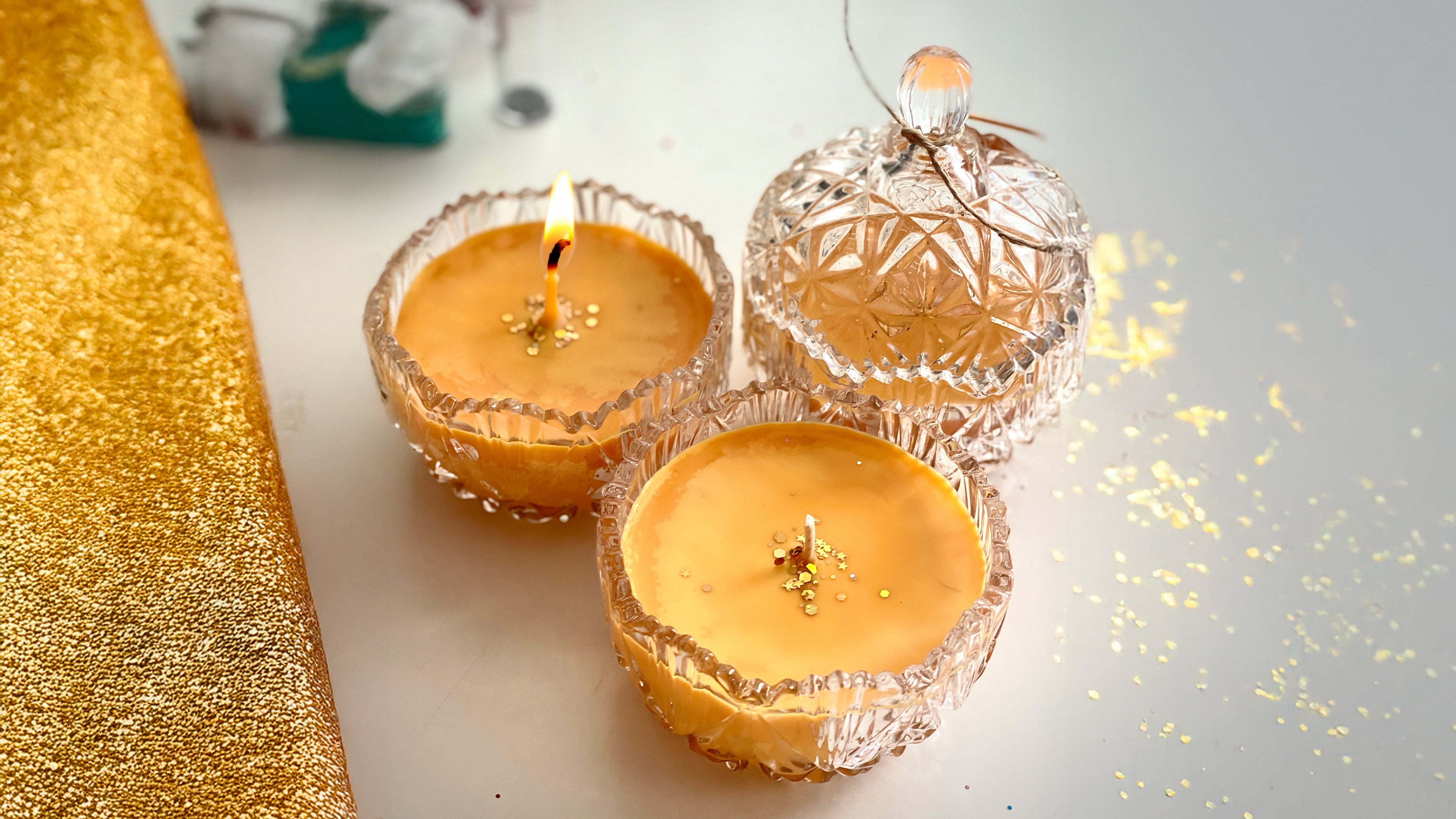 Three lit candles in decorative glass holders on a reflective surface with a gold glittery border.