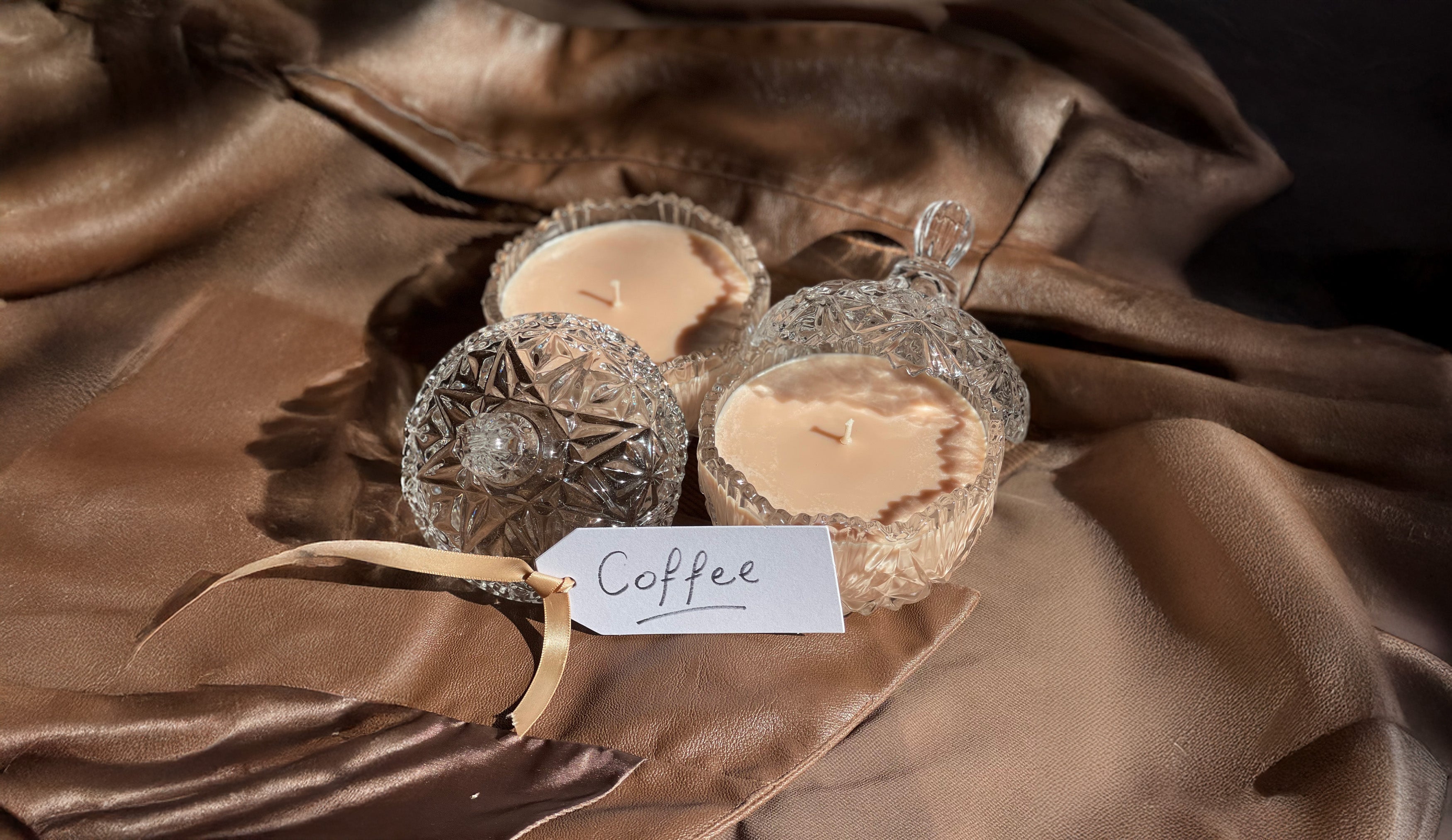 Decorative candles in glass holders with a 'Coffee' tag on a brown fabric background